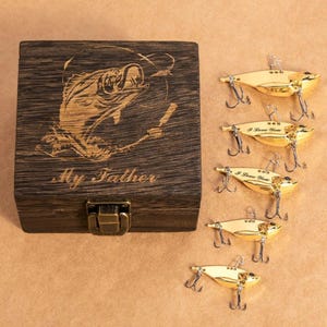 May include: A wooden box with a fish graphic and the text "My Father" is next to five gold-colored fishing lures. Each lure has three hooks and the text "I Love You". The box has a metal clasp.