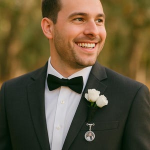 Può includere: Un uomo in abito grigio scuro con papillon nero e camicia bianca. Un'occhiello di rosa bianca è appuntato al bavero, insieme a un piccolo ciondolo rotondo con una foto. L'uomo sorride.