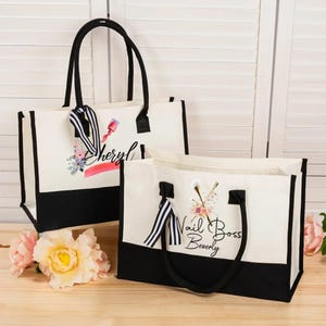May include: Two canvas tote bags with black handles and trim. The bags have a white upper section and a black lower section. One bag has the name "Sheryl" with nail polish art, and the other says "Nail Boss Beverly" with nail art.