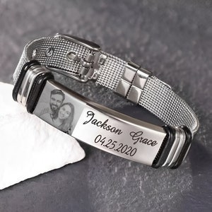 May include: A silver mesh bracelet with a buckle clasp. The bracelet features a rectangular plate with a photo of a couple and the engraved text "Jackson Grace 04.25.2020". The bracelet is on a white surface.