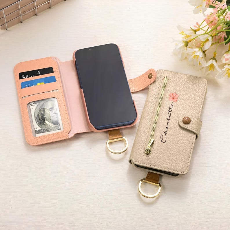 May include: A peach-colored phone case with card slots and a clear ID window, next to a beige wallet-style phone case with a zipper and the name "Charlotte" in script. Both cases have gold-colored hardware.