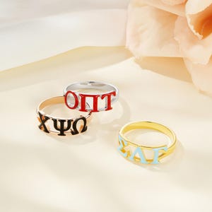 May include: Three rings with Greek letters in various colors. One ring is gold with light blue letters, another is gold with black letters, and the third is silver with red letters. The rings are displayed on a light-colored surface.