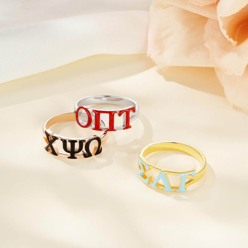May include: Three rings with Greek letters in various colors. One ring is gold with light blue letters, another is gold with black letters, and the third is silver with red letters. The rings are displayed on a light-colored surface.