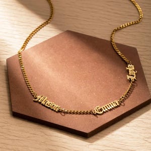 Pode incluir: Um colar dourado com três pingentes com nomes: "Alison", "Emma" e "Jade". Os nomes estão em uma fonte inglesa antiga. O colar tem um design de corrente de elos. O fundo é uma forma hexagonal marrom.