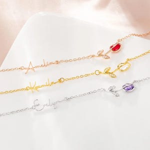 May include: Three delicate name necklaces in rose gold, gold, and silver. Each necklace features a cursive name and a rose pendant with a red, clear, or purple gemstone. The necklaces are displayed on a white surface.