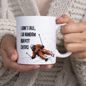 May include: A white ceramic mug featuring a graphic of a skier in an orange suit and helmet, holding ski poles while leaning on snow. The text printed in black reads, I DON'T FALL, I DO RANDOM GRAVITY CHECKS, positioned above the skier illustration.