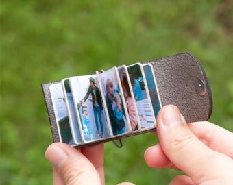 May include: A small, brown leather photo album with a snap closure, open to display several small photographs. The album is held in a person's hand against a blurred green background.