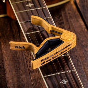 Op de afbeelding: Een houten gitaar capo met de naam "Nick" erop gegraveerd. De capo heeft ook de tekst "Keep on strumming, all my love Alexa xx" erop gegraveerd.