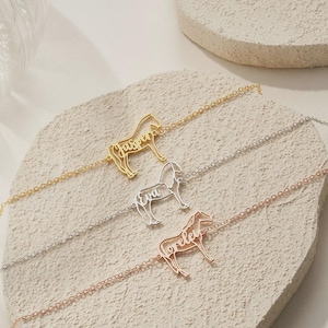 Puede incluir: Tres pulseras personalizadas con forma de caballo en oro, plata y oro rosa. Cada pulsera presenta un nombre: "Jasper", "Eva" y "Lorelei". Las pulseras se exhiben sobre una superficie texturizada de color claro.
