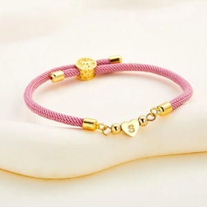 Puede incluir: Un brazalete de cordón trenzado rosa con un dije de corazón dorado y una letra 'S' dorada en el dije. El brazalete tiene un cierre dorado con un diseño decorativo.