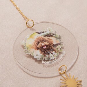 May include: A clear, circular ornament featuring a color photo of a brown dog surrounded by a floral wreath. The words "FOREVER LOVED" are printed on the ornament. A gold chain and sun charm are attached.