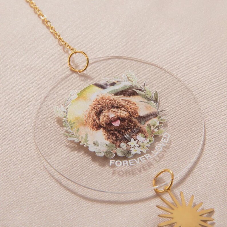 May include: A clear, circular ornament featuring a color photo of a brown dog surrounded by a floral wreath. The words "FOREVER LOVED" are printed on the ornament. A gold chain and sun charm are attached.