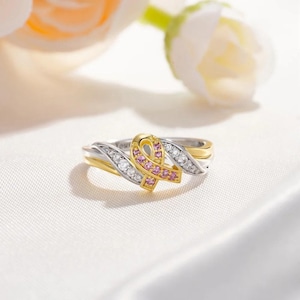 May include: A gold and silver ring with a pink ribbon design. The ribbon is set with small pink stones and the ring is decorated with clear stones.