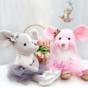 May include: Two plush toy mice, one gray and one pink, are featured in this image. The gray mouse wears a gray tutu and silver bow. The pink mouse wears a pink tutu and has the name "Ruby" on it. A small mouse illustration hangs above.