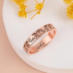 May include: Rose gold ring with a floral pattern etched around the band. The ring has the name "Sophia" engraved on the side. The ring is displayed on a white surface with yellow flowers in the background.