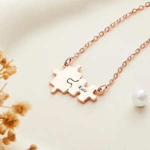 Pode incluir: Colar de ouro rosa com duas peças de quebra-cabeça que se encaixam. A peça da direita tem o nome "Kate" gravado.