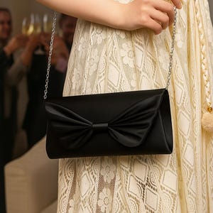 K&ouml;nnte beinhalten: Schwarze Satin-Clutch mit gro&szlig;er Schleife auf der Vorderseite. Die rechteckige Tasche hat einen silbernen Kettenriemen. Im Hintergrund ist eine Person in einem cremefarbenen Spitzenkleid zu sehen.