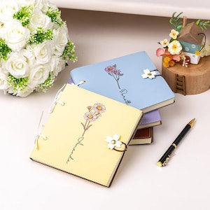 May include: A stack of floral-themed notebooks in pastel colors, including yellow and blue, with daisy illustrations and the names "Amanda" and "Dorath" printed on them. A black and gold pen rests nearby.