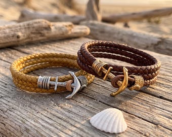 May include: Two braided leather bracelets, one mustard yellow and one dark brown, each featuring a metal anchor-shaped clasp. The yellow bracelet has a silver-toned anchor, while the brown bracelet has a gold-toned anchor. A small white seashell sits nearby.