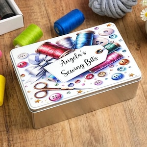 May include: A rectangular metal sewing box with a white lid featuring a colorful design of spools of thread, buttons, and a bow. The box has the text "Angela's Sewing Bits" on a tag. The box is on a wooden surface.