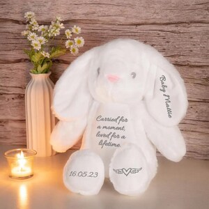May include: A white plush bunny with long ears, a pink nose, and gray embroidered text. The text on the bunny reads "Baby Mattie" and "Carried for a moment, loved for a lifetime." The bunny's feet have the date "16.05.23" and a heart with wings.