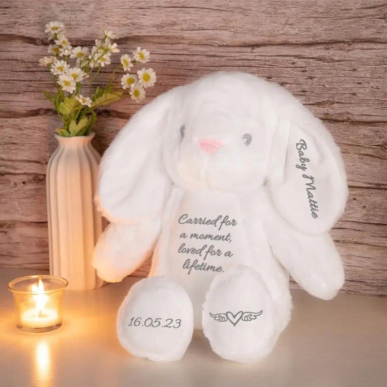 May include: A white plush bunny with long ears, a pink nose, and gray embroidered text. The text on the bunny reads "Baby Mattie" and "Carried for a moment, loved for a lifetime." The bunny's feet have the date "16.05.23" and a heart with wings.