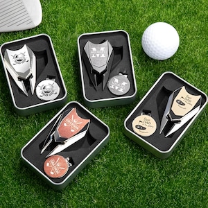 Könnte beinhalten: Fünf silberne Golfball-Markiersätze in individuellen Geschenkboxen. Jedes Set enthält einen silbernen Golfball-Marker mit einer personalisierten Inschrift und einen silbernen Golfball-Retriever. Die Inschriften beinhalten "Michael", "Grayson", "Bester Papa der Galaxie" und zwei mit dem Buchstaben "M".