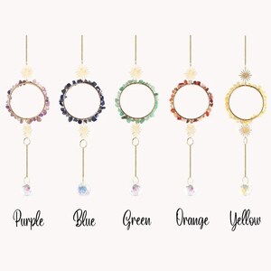 May include: Five hanging suncatchers with gemstone accents in purple, blue, green, orange, and yellow. Each features a gold-colored ring adorned with small stones, a starburst, a chain, and a crystal ball. The colors are labeled below each suncatcher.