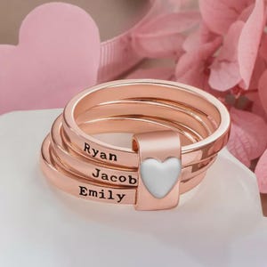 Pu&ograve; includere: Anelli impilabili in oro rosa con i nomi "Ryan", "Jacob" ed "Emily" incisi. Un dettaglio a forma di cuore bianco &egrave; sul lato. Gli anelli sono impilati, creando un effetto a strati.