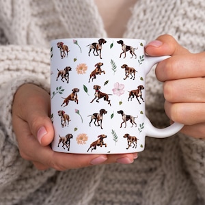 May include: White ceramic mug featuring a repeating pattern of brown and white dogs, flowers, and green leaves. The mug has a curved handle and is held by a person wearing a cream-colored sweater. The design is a watercolor style.