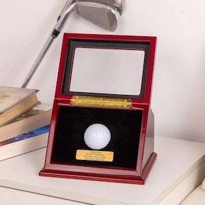 Könnte beinhalten: Ein roter, hölzerner Golfball-Präsentationskasten mit Glasdeckel. Der Kasten enthält einen weißen Golfball und eine Plakette mit der Aufschrift "Hole in one George Koutsoukos 4/12/2023 Hole # 140m Warrensburg Golf Club".