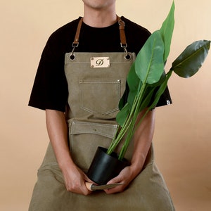 Peut inclure: Une personne portant un tablier en toile marron avec une sangle en cuir et une boucle en métal. Le tablier a une grande poche sur le devant et une petite étiquette en métal avec la lettre "D" dessus. La personne tient une plante en pot avec des feuilles vertes.