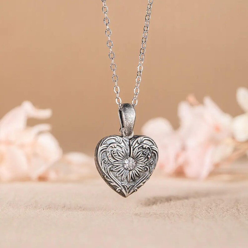 May include: A silver heart-shaped locket necklace with an ornate floral design and a small clear rhinestone in the center.