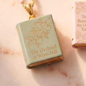 May include: A sage green book-shaped pendant necklace featuring a gold-toned metal bail and a gold-engraved illustration of an apple branch. The cover displays the text The Orchard at Wren Hill in gold lettering, with gold-toned page edges visible.