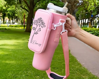 May include: Pink insulated water bottle carrier with a black floral design and the word "Dreamy" printed on it. The carrier has a shoulder strap and a small pocket.