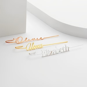 May include: Three personalised hair barrettes with the names "Olivia", "Alexa", and "Elizabeth" written in cursive. The barrettes are made of metal and are available in gold, rose gold, and silver.