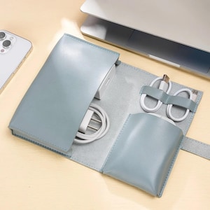 Könnte beinhalten: Ein hellblaues Leder-Tech-Organizer, geöffnet, um ein weißes Netzteil und Kabel freizugeben. Zwei weiße Ladekabel sind mit grauen Riemen befestigt. Eine kleine Tasche ist ebenfalls sichtbar. Ein Smartphone und ein Laptop befinden sich im Hintergrund.
