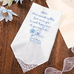 Op de afbeelding: Witte zakdoek met blauw geborduurde tekst en bloemmotief. De tekst luidt: "Alida, Hier is iets blauws van mij voor jou om gelukkige tranen op te vangen als je 'ja' zegt." Bevat de datum "10-05-2024".