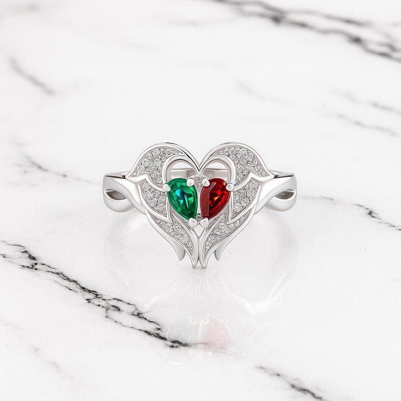 Puede incluir: Anillo de plata con dise&ntilde;o de coraz&oacute;n con dos gemas en forma de pera, una verde y otra roja. El coraz&oacute;n est&aacute; adornado con peque&ntilde;as piedras brillantes. Fondo de m&aacute;rmol blanco.