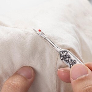 A metal tool with a sharp tip and a hook that can cut threads and seams. The tool can be customized with a name engraved on the handle. It is a vintage style sewing gift for tailors, seamstresses, or fashion designers.