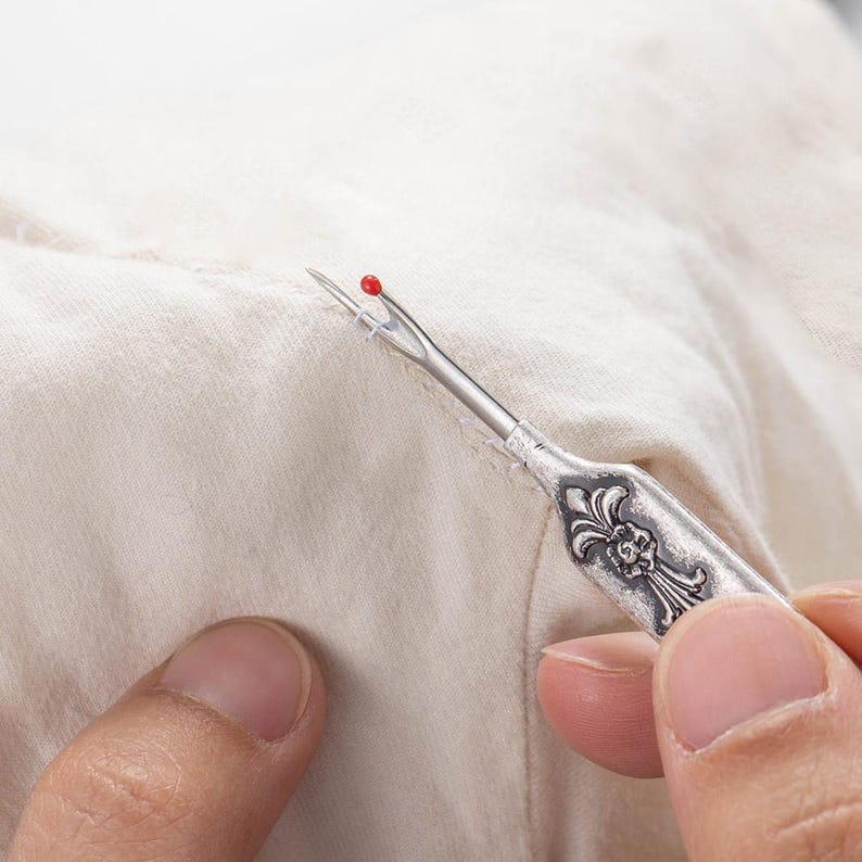 A metal tool with a sharp tip and a hook that can cut threads and seams. The tool can be customized with a name engraved on the handle. It is a vintage style sewing gift for tailors, seamstresses, or fashion designers.