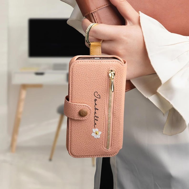 May include: A pink leather phone case with a gold zipper and a snap closure. The case has the name "Isabella" written on it in cursive, along with a small white flower. A gold ring is attached to the top.