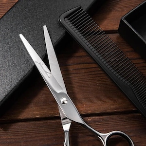 May include: A pair of silver hairdressing shears with the word "Jonathan" engraved on the blade. The shears are lying on a wooden surface next to a black comb.