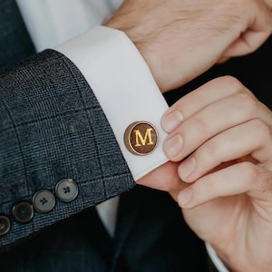May include: Close-up of a pair of wooden cufflinks with a gold-coloured border and the letter "M" in gold. The cufflinks are on a white dress shirt cuff, paired with a grey plaid suit jacket.