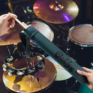 May include: A dark green drumstick case with the brand name "Oakley" in a script font. The case is held by a person's hands, with drumsticks partially visible. The background features a drum set with cymbals and a tambourine.