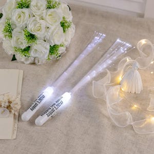 Può includere: Due bacchette bianche in fibra ottica con la scritta "Happily Ever Austin August 5, 2024#" sono esposte. Un bouquet di rose bianche e un piccolo vestito decorativo sono sullo sfondo. Le bacchette emettono una luce bianca soffusa, perfetta per un matrimonio.