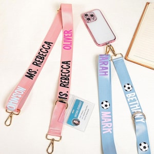 Puede incluir: Dos cordones, uno rosa y otro azul claro, con cierres dorados. El cordón rosa tiene el texto "MS. REBECCA JOHNSON" y "OLIVER". El cordón azul claro tiene los nombres "SARAH", "MARK" y "BETHANY" en rosa, con gráficos de balones de fútbol.