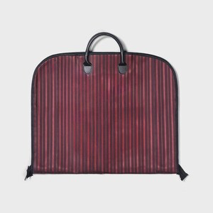 Puede incluir: Una funda de ropa burdeos con rayas verticales en diferentes tonos de rojo. La bolsa tiene una cremallera negra, ribete negro y un asa negra con dos agarres. La bolsa est&aacute; dise&ntilde;ada para transportar ropa.