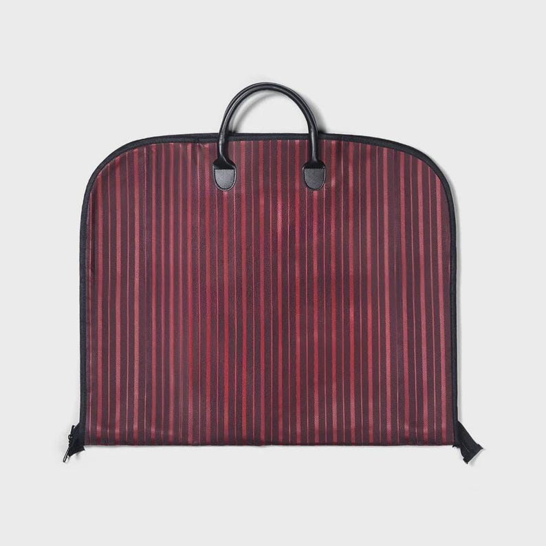 Puede incluir: Una funda de ropa burdeos con rayas verticales en diferentes tonos de rojo. La bolsa tiene una cremallera negra, ribete negro y un asa negra con dos agarres. La bolsa est&aacute; dise&ntilde;ada para transportar ropa.
