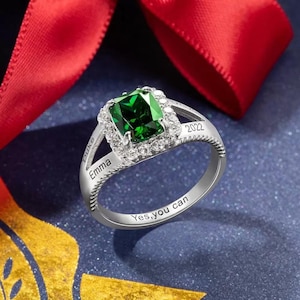 Puede incluir: Un anillo de plata con una piedra preciosa verde de corte esmeralda rodeada de piedras más pequeñas y transparentes. El anillo está grabado con las palabras "Duke", "Emma", "2022" y "Yes, you can".